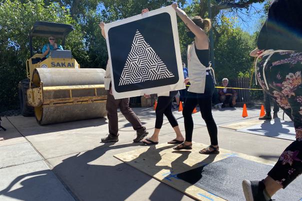 People carry a large black-and-white geometric print past a steamroller outdoors.