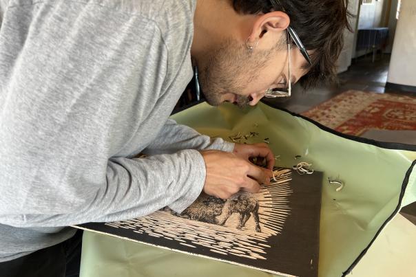 Artist Jacob Gutierrez leans over a work table, carving a detailed woodcut block with hand tools inside the Risolana studio.