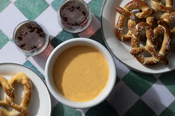Bowl of golden beer-cheese dip on a checkered table with soft pretzels and two dark beers.