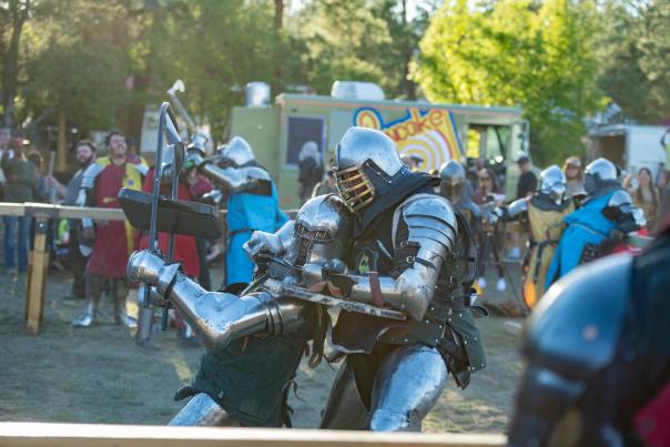 Two armored knights engage in intense combat at an outdoor medieval festival, with onlookers watching and a colorful food truck in the background.