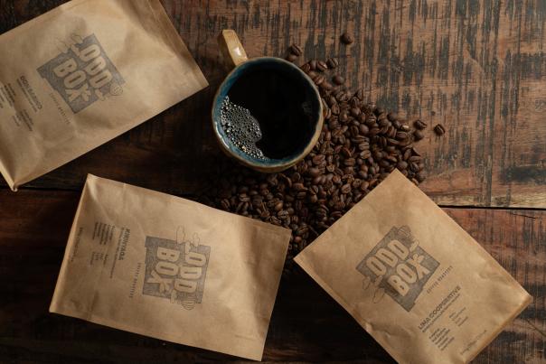 A cup of coffee sits on a wooden table surrounded by roasted coffee beans and three bags of "Odd Box Coffee Roasters" coffee beans.