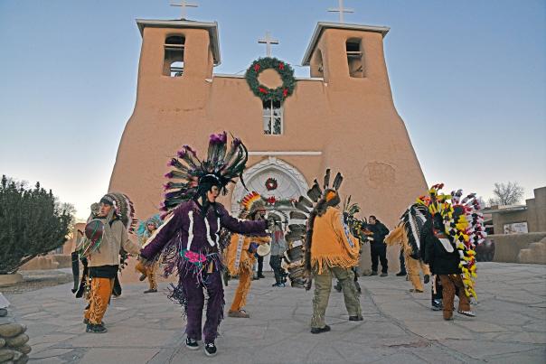 A group of Indigenous dancers wearing colorful feathered headdresses and traditional attire perform in front of a historic adobe church with wreaths, beneath a clear sky.
