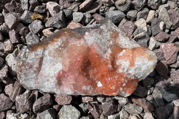 A geode of clear and reddish-orange quartz crystals rests on a bed of gray and brown rocks.