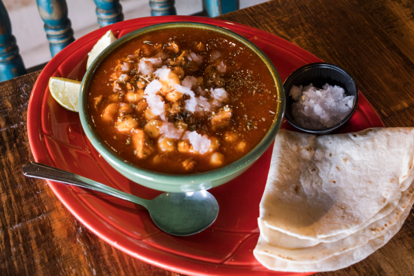A bowl of red posole sits on a red plate with a side of corn tortillas and chopped white onion.