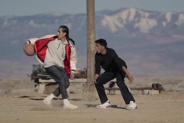Two young men play basketball on a dusty court with mountains in the distance.