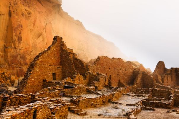 The remains of stone buildings, part of ancient ruins, stand in front of a tall, sheer cliff face.