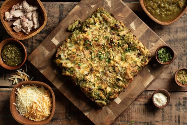 Square green chile pizza on a wooden board surrounded by bowls of cheese, chicken, pesto, chiles, and herbs.