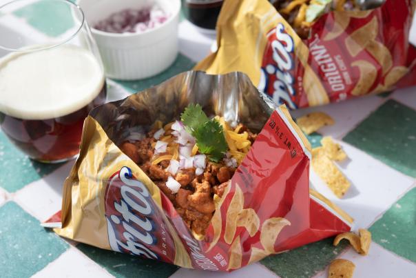 Two open Frito pie bags filled with red-chile meat, beans, cheese, and onions with a pint of beer nearby.