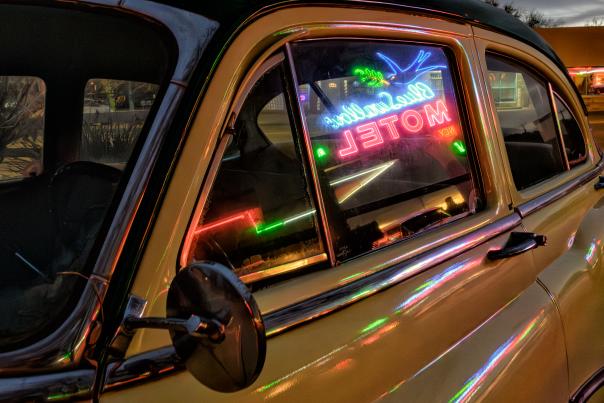 A classic car reflects the neon glow of a motel sign.