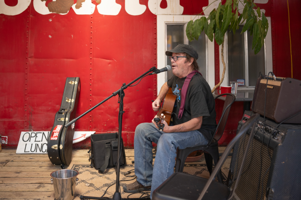 A man sits on a chair playing an acoustic guitar in front of a red wall with "Santa Fe" painted on it.
