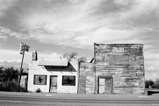 A black and white photo of two dilapidated buildings, one brick and one wood, with boarded up windows and a crooked sign, stand on a roadside.