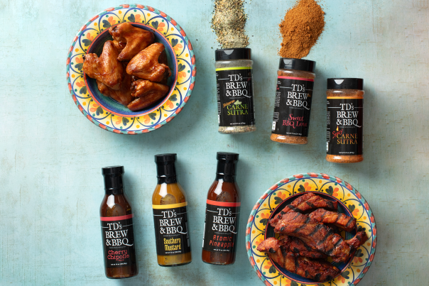 A flat lay display shows colorful plates of cooked chicken wings and ribs surrounded by TD’s Brew & BBQ seasoning bottles and sauces arranged on a light blue surface.