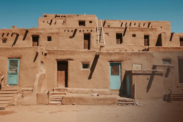 A multi‑story adobe dwelling with tan earthen walls, wooden doors, small square windows, and ladders stands under a clear blue sky.