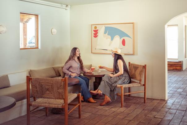 Two women sit at a round table in a cozy, minimalist room with ceramic floor tiles.
