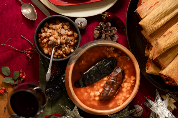 A festive table with traditional Mexican dishes, including pozole, tamales, and a glass of red wine, with holiday decorations scattered around.