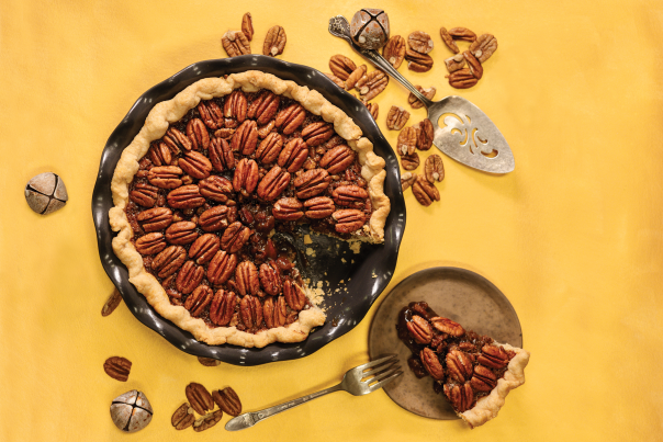 A pecan pie with one slice removed sits on a yellow tablecloth with pecans and jingle bells scattered around.