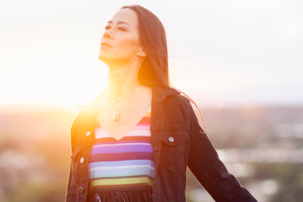 A woman in a black jacket and a colorful dress leans back against a railing with her eyes closed as the sun sets behind her.