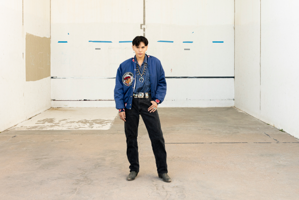 Jojo Jackson in a blue jacket with a beaded design, black jeans, and boots stands in a concrete room.