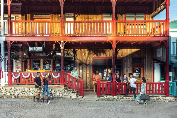 Two-story wooden building with red railings. People gather outside under warm sunlight; a couple walks two dogs, and others chat casually.