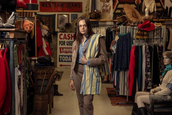 A young adult stands in a vintage clothing store, surrounded by racks of clothes and mannequins.