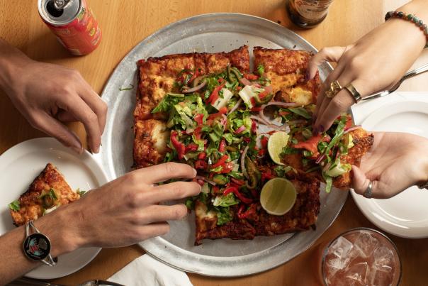 Hands grabbing slices of a rectangular pizza topped with fresh vegetables and lime wedges on a metal tray.