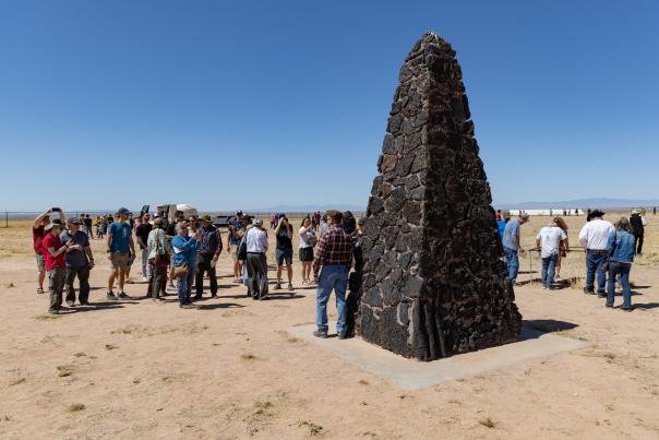 Group of visitors gathered around a tall stone monument in a desert landscape under a clear sky.