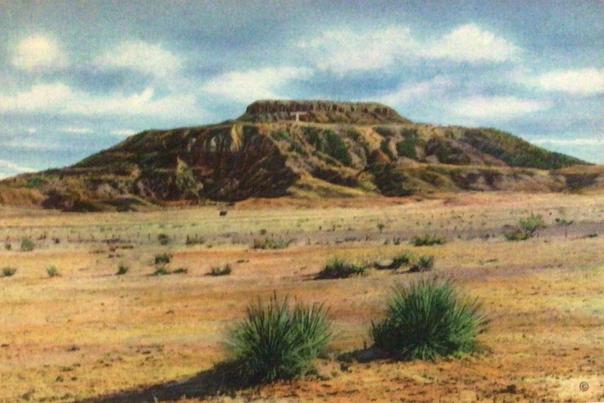 A flat-topped, rocky mesa rises above a dry desert plain dotted with low shrubs under a partly cloudy sky.