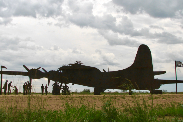 A silhouette of a bomber plane sits on a grassy field with a line of people figures standing nearby.