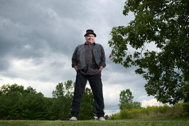 A man stands confidently in a park, hands in his pockets, wearing a black vest and hat.