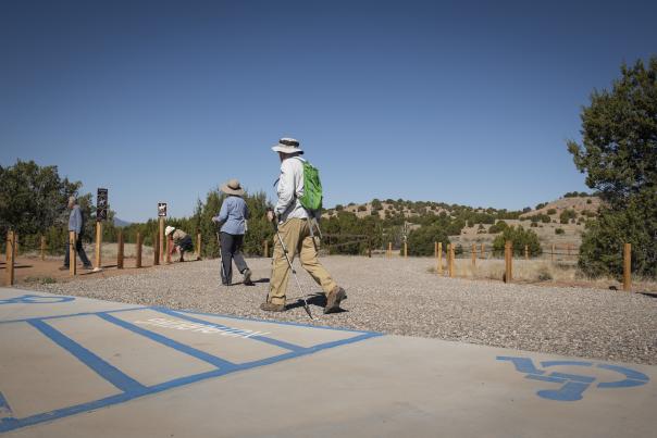 People with hats and backpacks walk along a sunny trailhead in a desert landscape with scattered trees and hills.