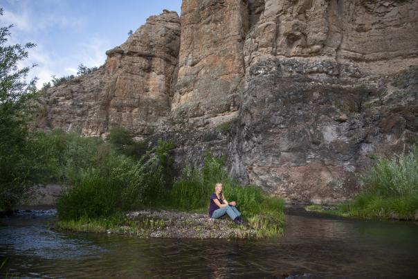 Person sits on a grassy riverbank surrounded by tall cliffs and flowing water.