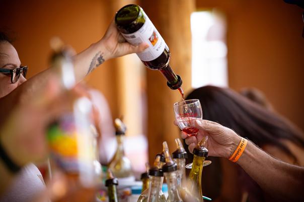 Person pours red wine from a bottle into a small glass at a booth lined with various bottles.