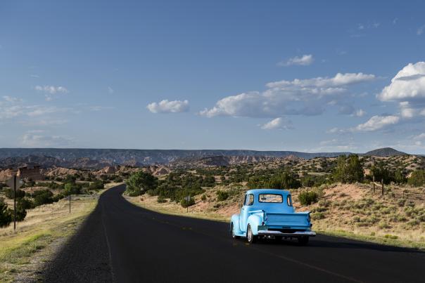 Vintage blue pickup drives a two-lane road of the High Road through sunlit badlands toward distant mesas.