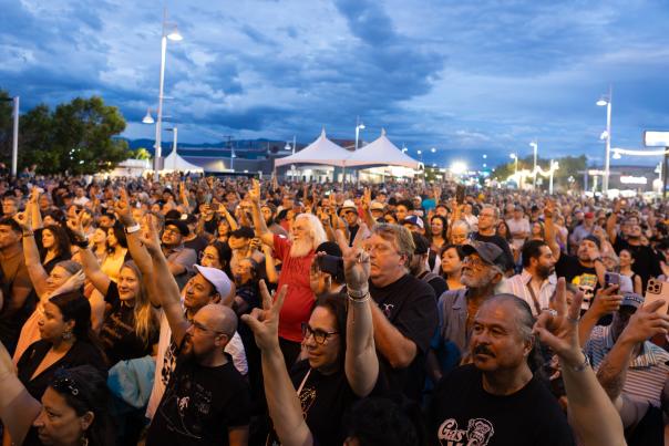 A large crowd at an outdoor event raises hands and phones toward a stage at dusk, with tents, lights, and a cloudy blue sky in the background.
