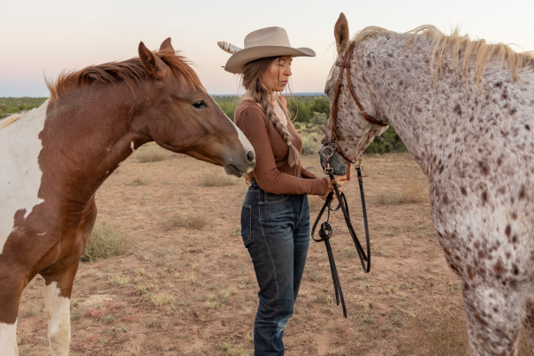 Artist Stella Maria Baer stands between two horses in the Santa Fe countryside, wearing a hat and holding a rope.