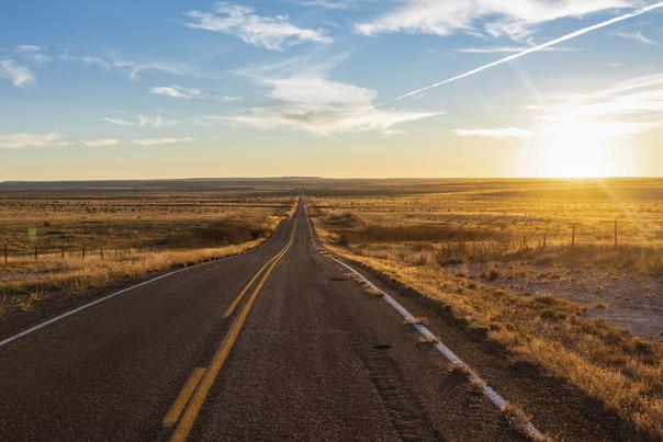 A two-lane road stretches toward the horizon through golden grassland beneath a blue and yellow sky.