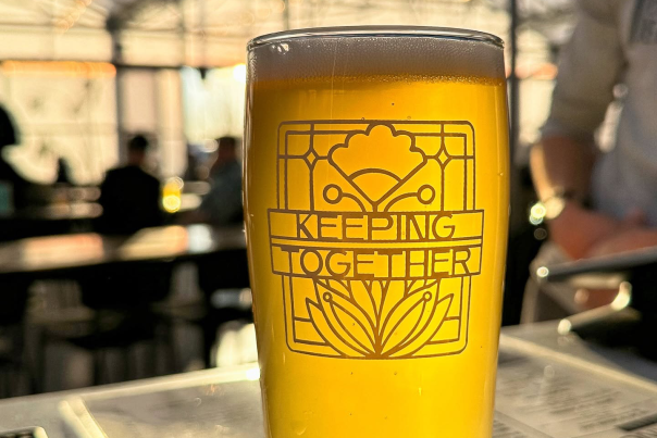 A tall glass of golden beer with a decorative ‘Keeping Together’ design is shown in close‑up on a bar counter with a softly lit taproom in the background.