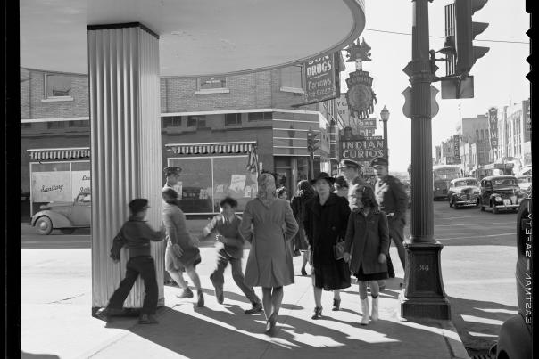 A lively 1940s street scene with people walking past a column and under a circular awning. Children play energetically. Vintage cars and storefronts line the background.