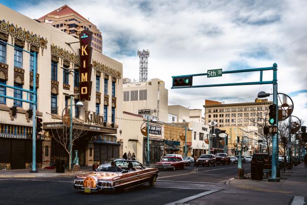 A colorful street scene shows classic cars driving past the ornate KiMo Theatre and surrounding buildings at the intersection of 5th Street in a busy downtown area.