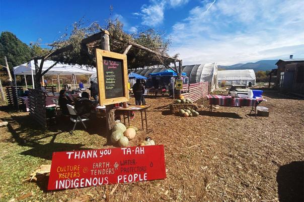 Outdoor market scene with a rustic wooden shelter adorned with greenery, tables of produce, and a red sign expressing gratitude to Indigenous people.