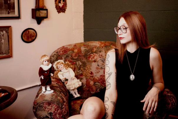A woman with glasses, tattoos, and a black dress sits in a vintage chair with two porcelain dolls in the background.