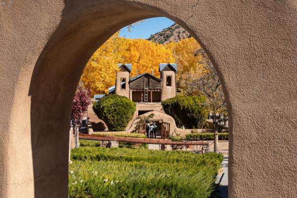 A rustic adobe arch topped with a simple cross frames a distant adobe chapel surrounded by golden autumn trees under a clear blue sky.