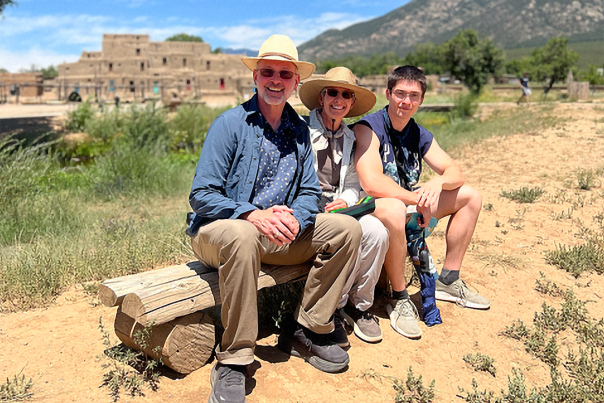 Three people sit smiling on a wooden bench outdoors on a sunny day. Behind them, a historic adobe structure and distant mountains under a clear blue sky.