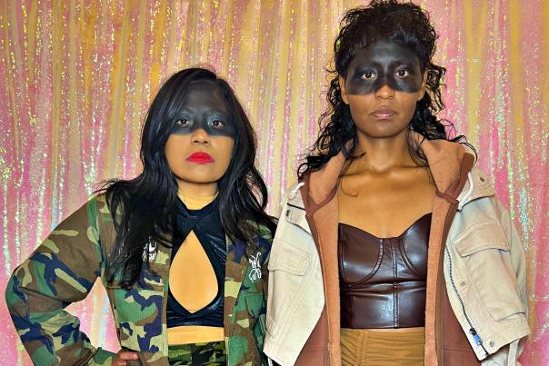 Sisters Natasha and Dorothy Cuylear, members of Lindy Vision, stand together in bold stage outfits, posing against a shimmering pink backdrop at a photo shoot.