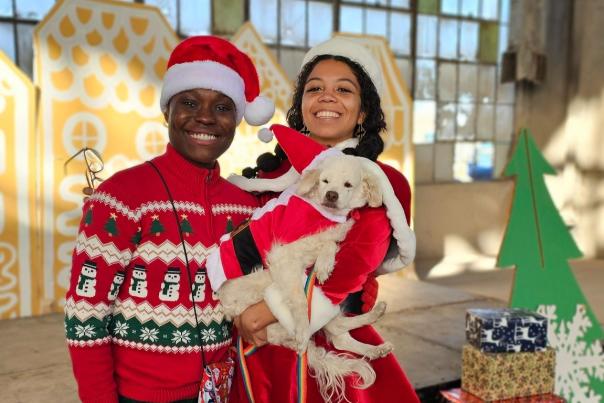 Two people in festive attire pose joyfully with a white dog in Santa costume. Background includes holiday decorations, presents, and a green tree.