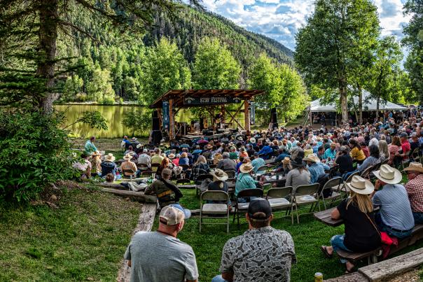 Outdoor music festival with a crowd gathered before a small lakeside stage in a forested setting.