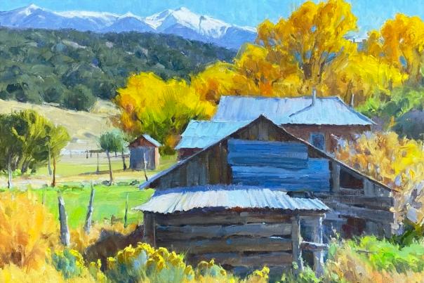 Rustic wooden barn with a tin roof set amidst vibrant autumn foliage. Snow-capped mountains and a clear blue sky in the background.