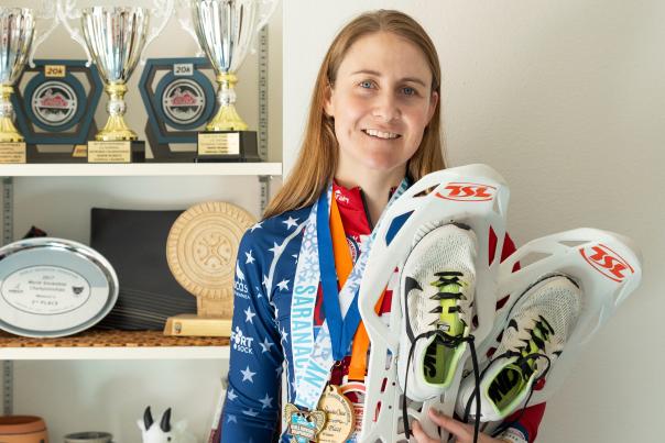 Michelle Hummel stands in front of her trophy shelf holding racing snowshoes and wearing medals.