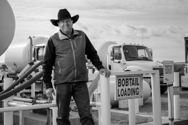 A person in a hat and outdoor work clothing stands beside a “Bobtail Loading” sign at a propane loading area with trucks and equipment in the background.