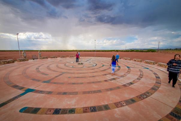 People walk on a circular labyrinth on a concrete surface under a dramatic, cloudy sky.